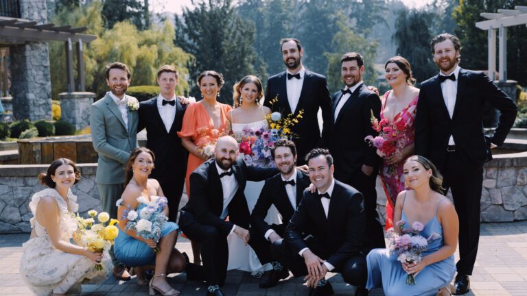 A wedding party of eleven people dressed in formal attire posing for a photo in Victoria, BC, holding colorful bouquets, with trees and a clear sky in the background.