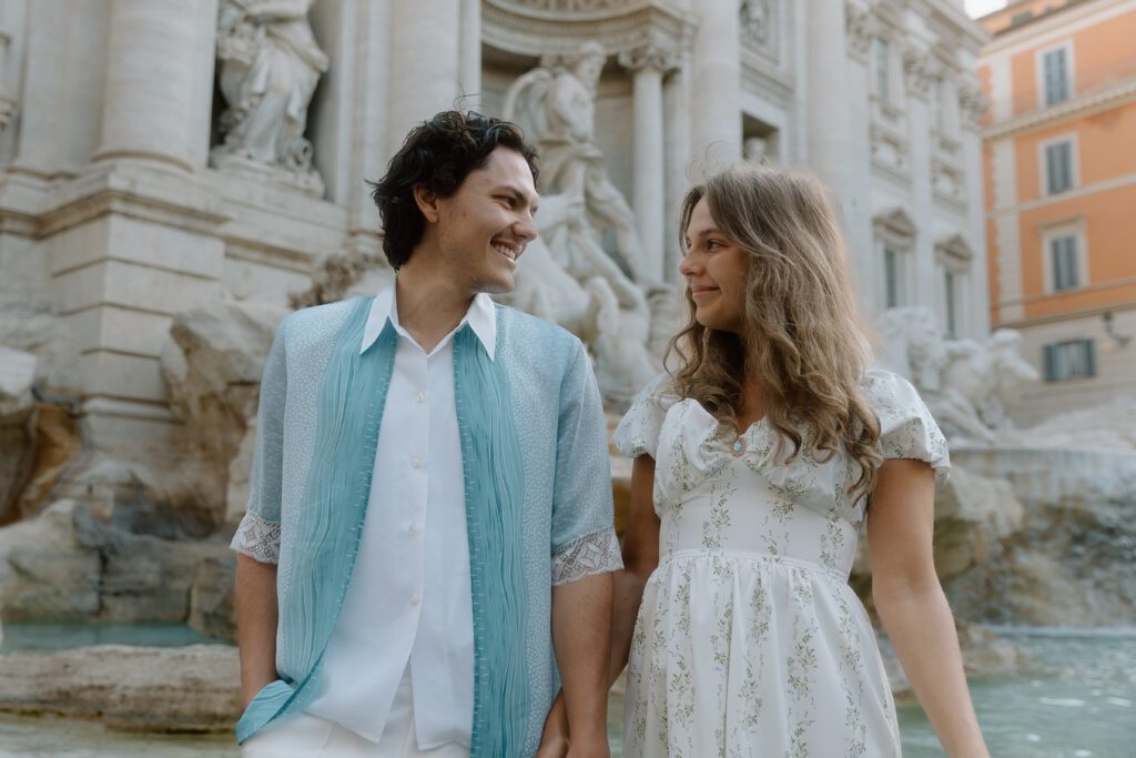 A couple look at each other smiling with Rome's Trevi Mountain behind them. The woman wears a white dress with green flowers. The man wears a white shirt and blue outer shirt.