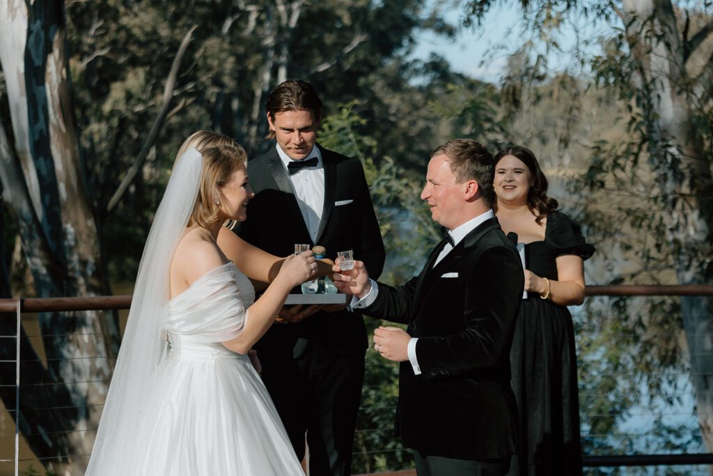 A bride and groom are exchanging small glass cups in an outdoor wedding ceremony. The bride is in a white gown and veil, while the groom is in a black suit. A female celebrant in black and one groomsman also in black, stand behind them in front of a wooded background, capturing the unity of their moment.