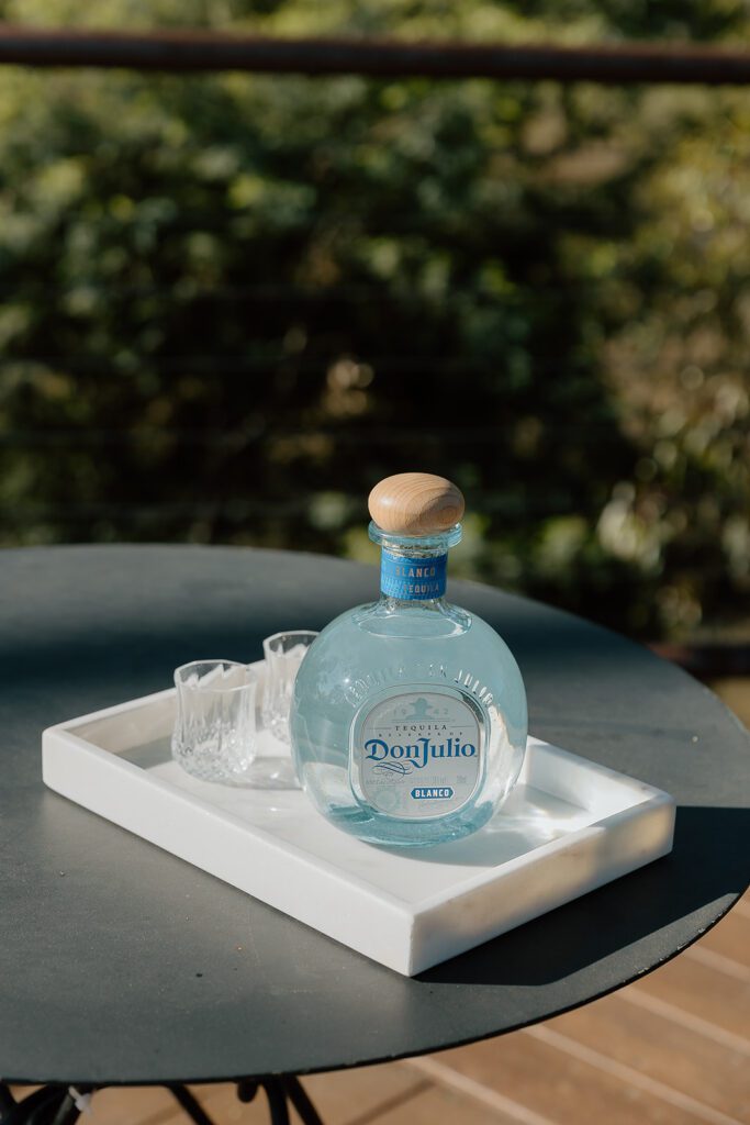 Bottle of Don Julio Tequila and two shot glasses on a black table, outside in the sun with greenery in the background.