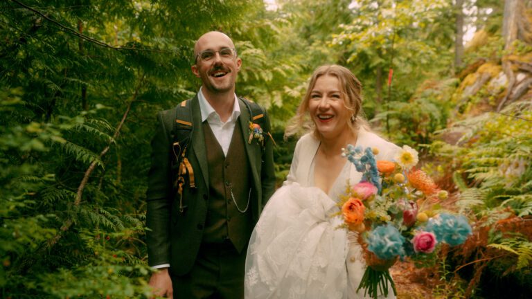 A groom in a green suit jacket with a black backpack smiles at the camera while the bride beside him wears a white dress, carries her train, and holds a bright, colourful bouquet, standing together in a lush forest on an overcast day in Squamish, British Columbia.
